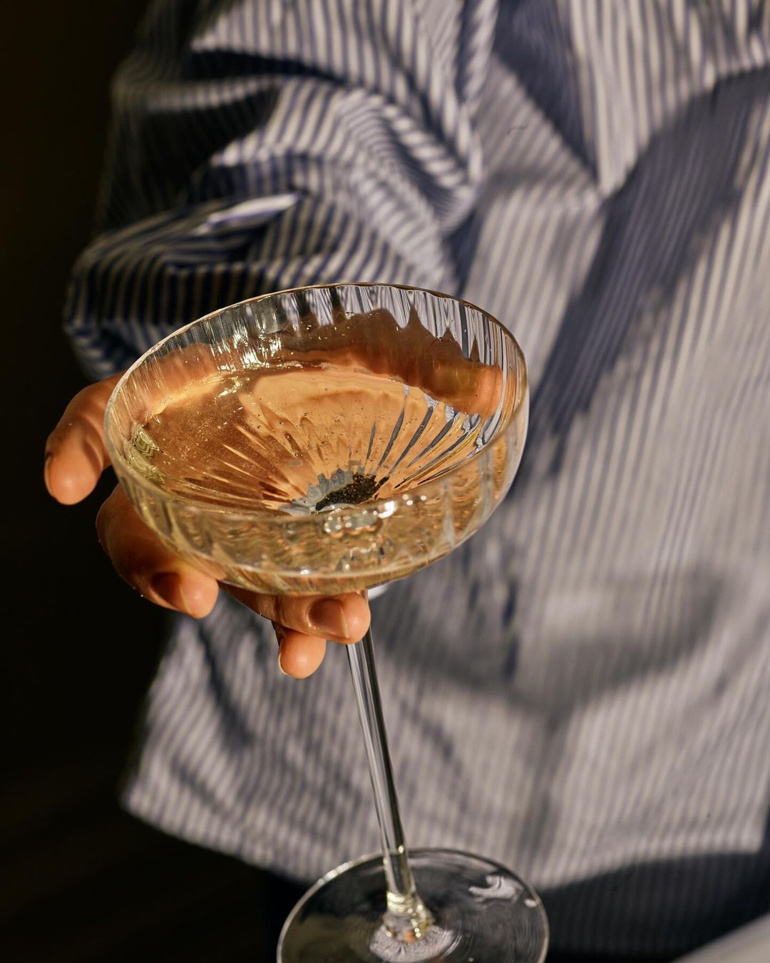 Crystal coupe glass with liquid held by a person wearing a blue and white striped shirt, close-up indoor shot, shallow depth of field.