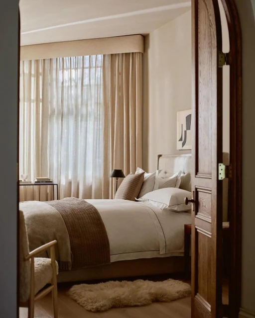 Bedroom interior showcasing a bed with beige and white linens, alongside a chair, through an arched wooden doorway, indoor lighting.