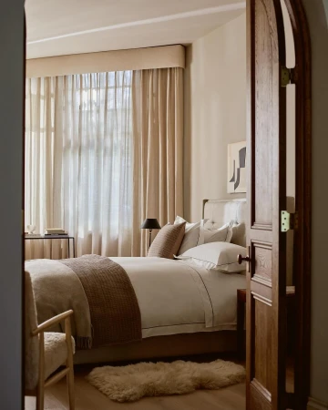 Bedroom interior showcasing a bed with beige and white linens, alongside a chair, through an arched wooden doorway, indoor lighting.