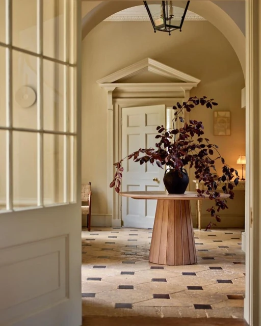 Interior view of a light beige hallway featuring a round wooden table with a dark floral arrangement, stone tile flooring, and arched doorways.