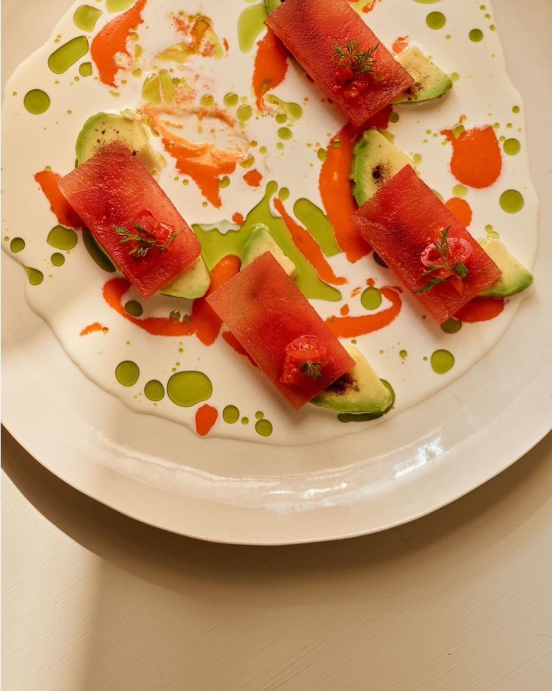 Gourmet food presentation featuring watermelon rolls with avocado slices arranged artistically on a white plate with decorative sauce.