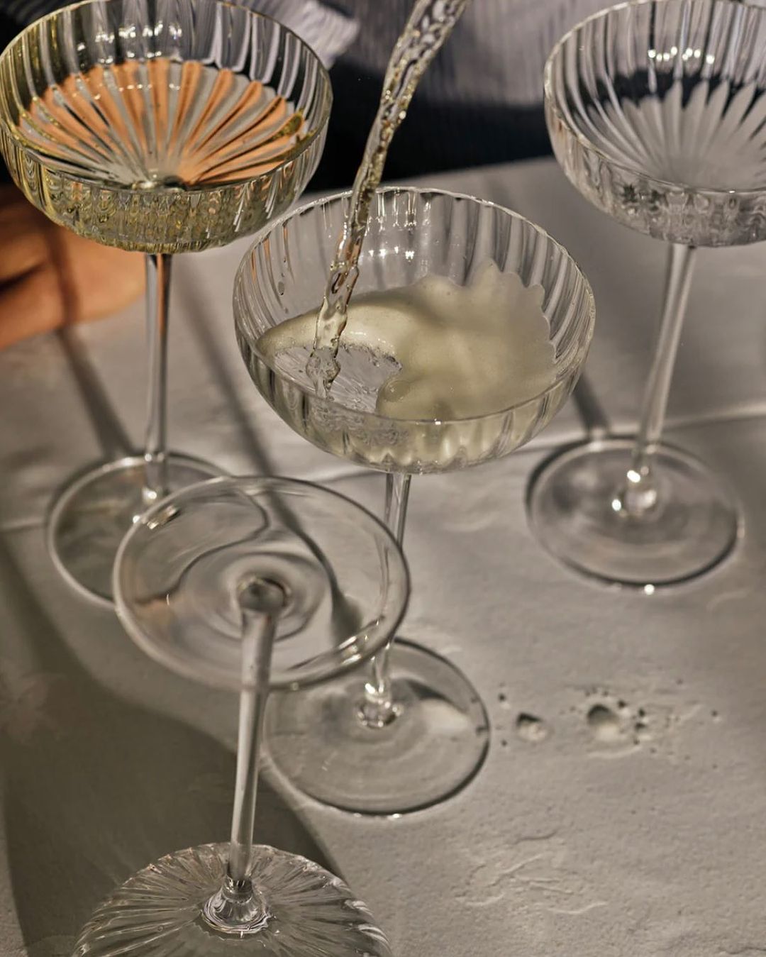 Champagne being poured into crystal coupe glasses on a white tablecloth, casting soft shadows in an indoor setting.