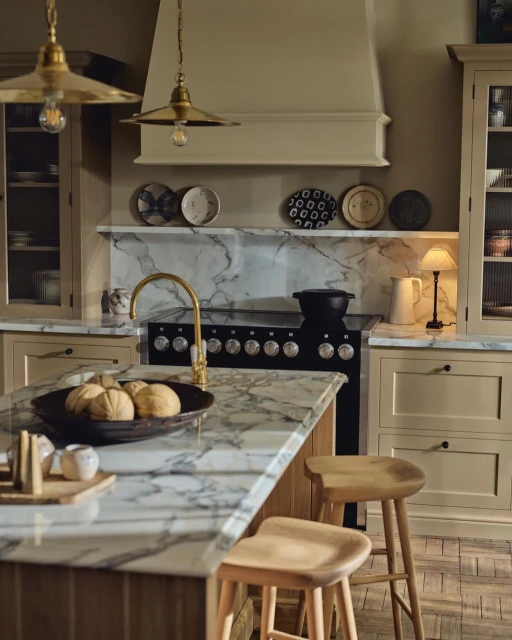 Elegant kitchen interior featuring marble countertops, light wood cabinets, a central island, and brass pendant lights, creating a warm ambiance.