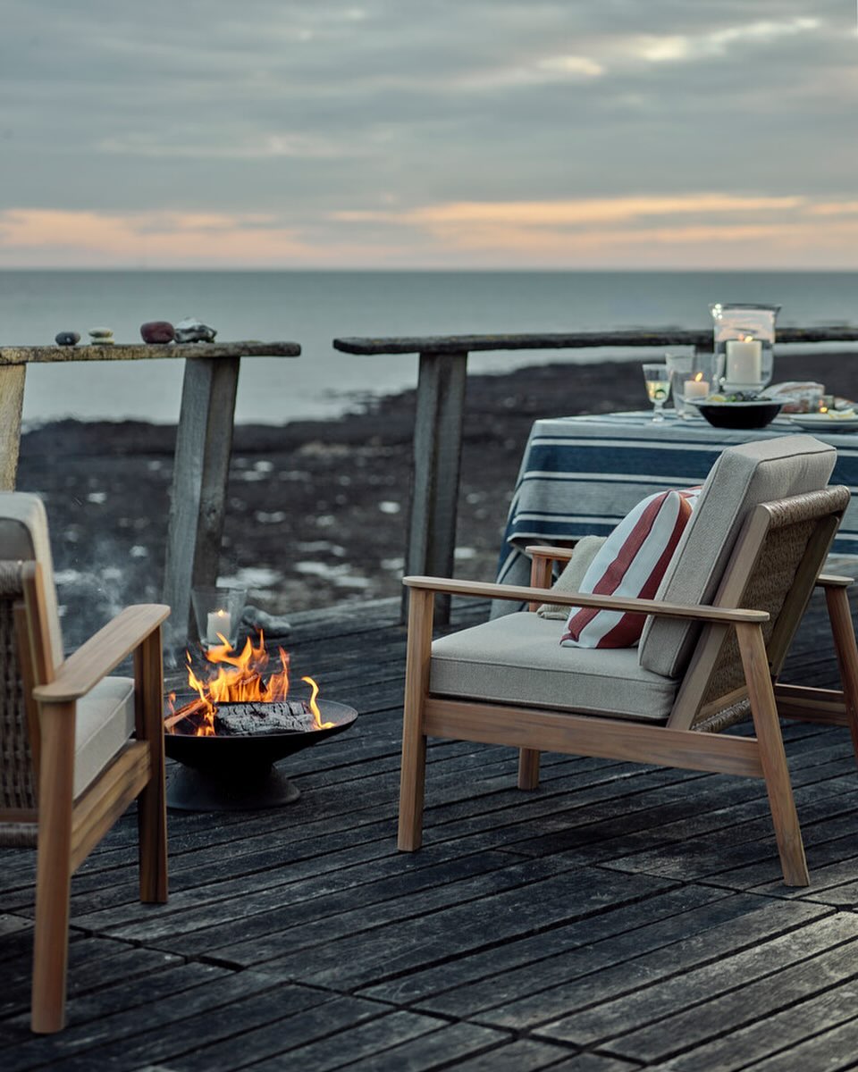 Outdoor patio scene features two wooden chairs, a fire pit with burning logs, a table with dishes, and a view of the ocean at dusk.