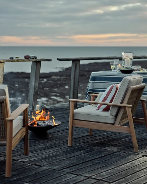 Outdoor patio scene features two wooden chairs, a fire pit with burning logs, a table with dishes, and a view of the ocean at dusk.