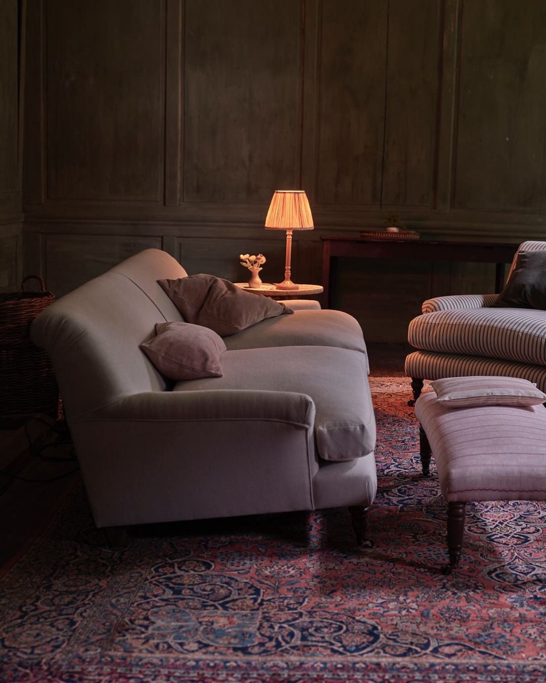 Living room interior with sofa, armchair, ottoman, table lamp, and patterned rug on hardwood flooring, set against dark green walls.