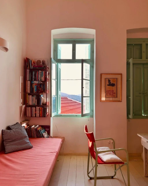 Living room interior with a daybed, books, and a window overlooking a red-tiled roof and mountains, creating a cozy and inviting atmosphere.