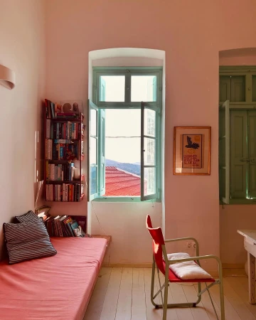 Living room interior with a daybed, books, and a window overlooking a red-tiled roof and mountains, creating a cozy and inviting atmosphere.