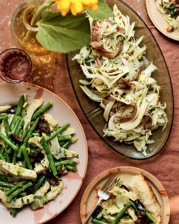 Overhead shot of arranged plates and platters featuring fresh salads with green beans, artichokes, fennel, and anchovies on a table.