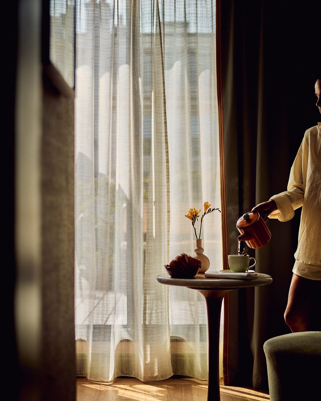Woman in neutral tones pours tea in a sunlit interior by window with sheer drapes, side table with flowers and pastry, and dark accents.