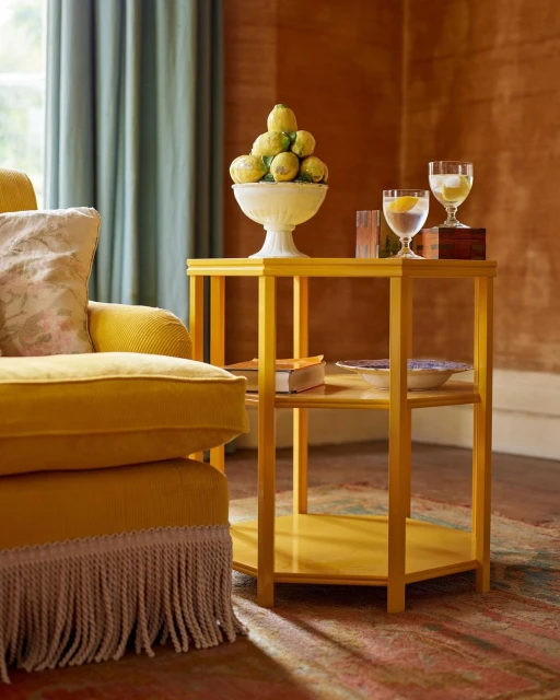Still life composition featuring a yellow side table with lemons in a white bowl, glasses, and a glimpse of a vintage interior.
