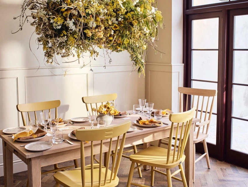 Dining room scene with a wooden table set for a meal, surrounded by wooden chairs. A floral arrangement hangs overhead, providing a soft light.