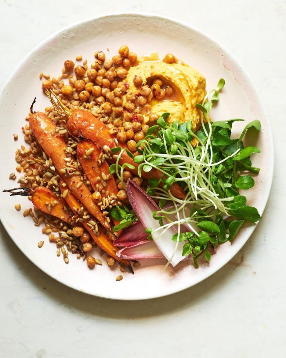 Overhead shot of a vibrant plate with roasted carrots, hummus, chickpeas, sprouts, and greens, set against a bright, clean white surface.