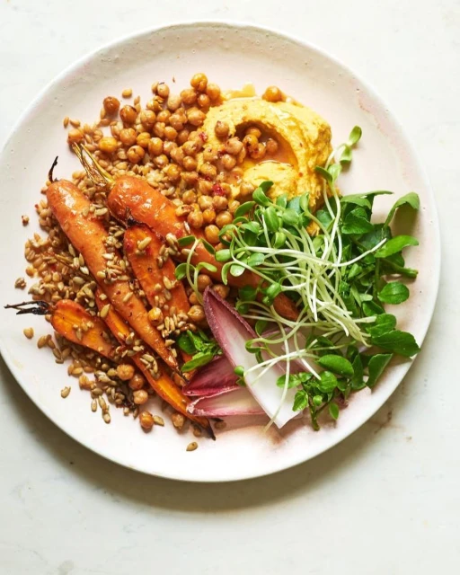 Overhead shot of a vibrant plate with roasted carrots, hummus, chickpeas, sprouts, and greens, set against a bright, clean white surface.