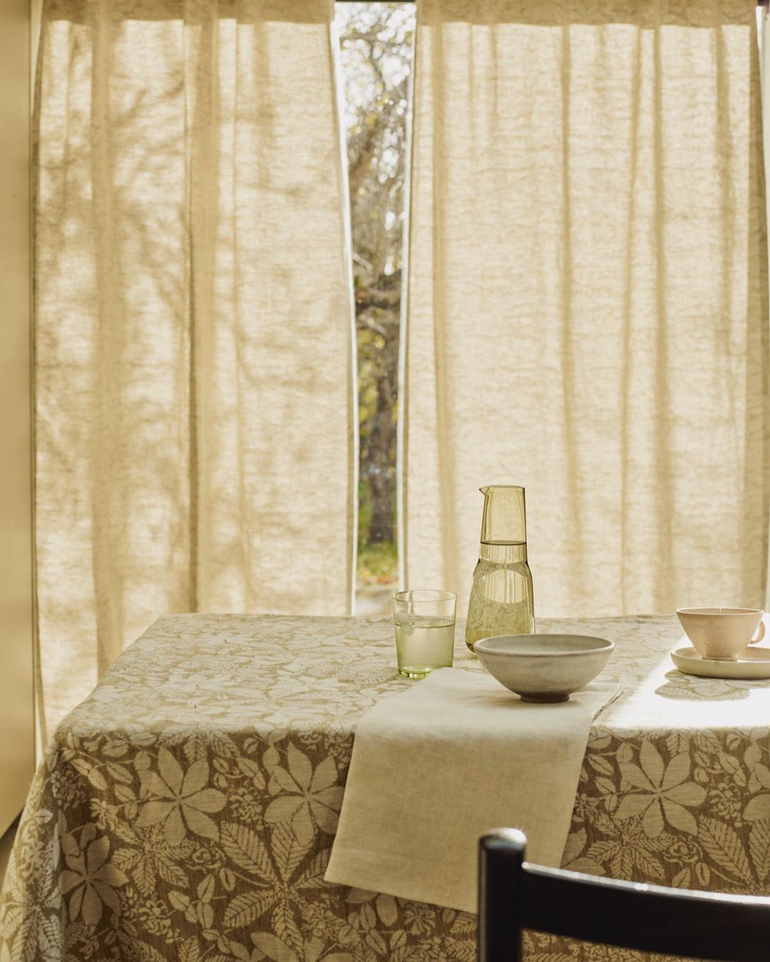 A dining table with a patterned tablecloth, a bowl, a pitcher, a glass, and a cup, set near a window with linen curtains.