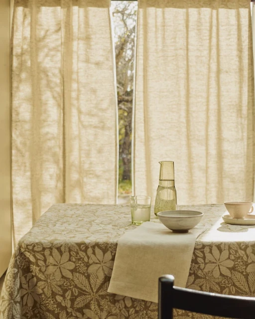 A dining table with a patterned tablecloth, a bowl, a pitcher, a glass, and a cup, set near a window with linen curtains.