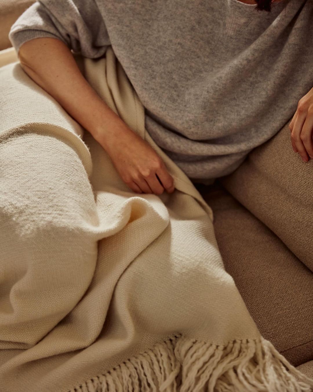 A woman is sitting on a beige couch and holding a soft, cream-colored fringed blanket, wrapped in a grey sweater and beige pants.