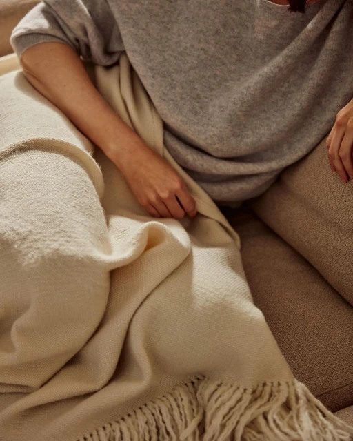A woman is sitting on a beige couch and holding a soft, cream-colored fringed blanket, wrapped in a grey sweater and beige pants.