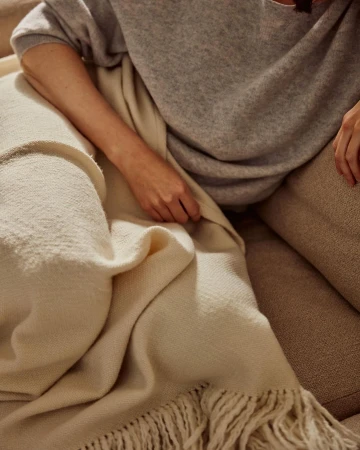 A woman is sitting on a beige couch and holding a soft, cream-colored fringed blanket, wrapped in a grey sweater and beige pants.