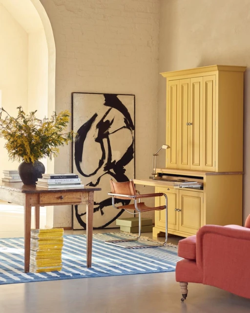 Living space showcases a wood desk with flowers, abstract art on the wall, yellow bureau, and a coral colored sofa for a serene atmosphere.