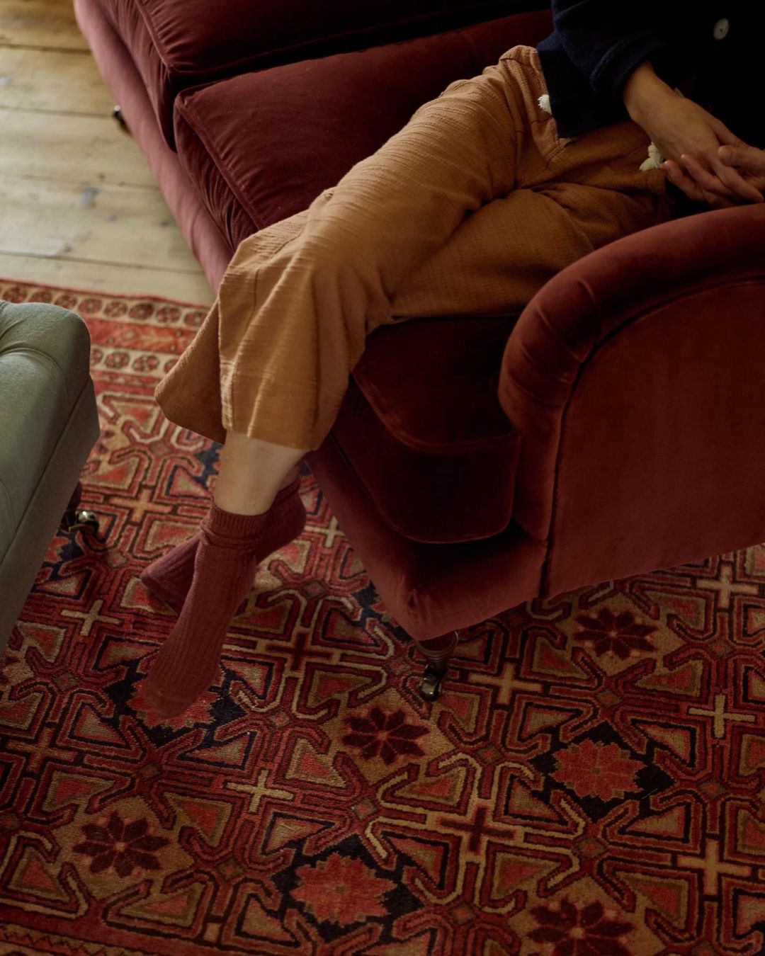 Person relaxing in a red velvet armchair in an indoor setting with patterned rug. The individual wears rust-colored pants and socks.