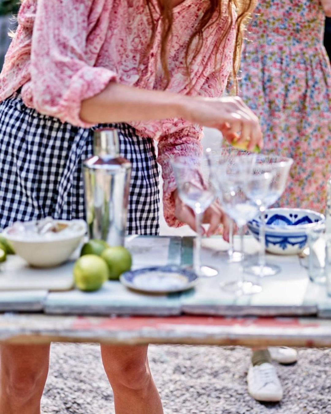 Two women prepare drinks at an outdoor bar with limes, glassware, and a shaker on a rustic table, enjoying a sunny day.