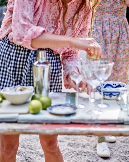 Two women prepare drinks at an outdoor bar with limes, glassware, and a shaker on a rustic table, enjoying a sunny day.