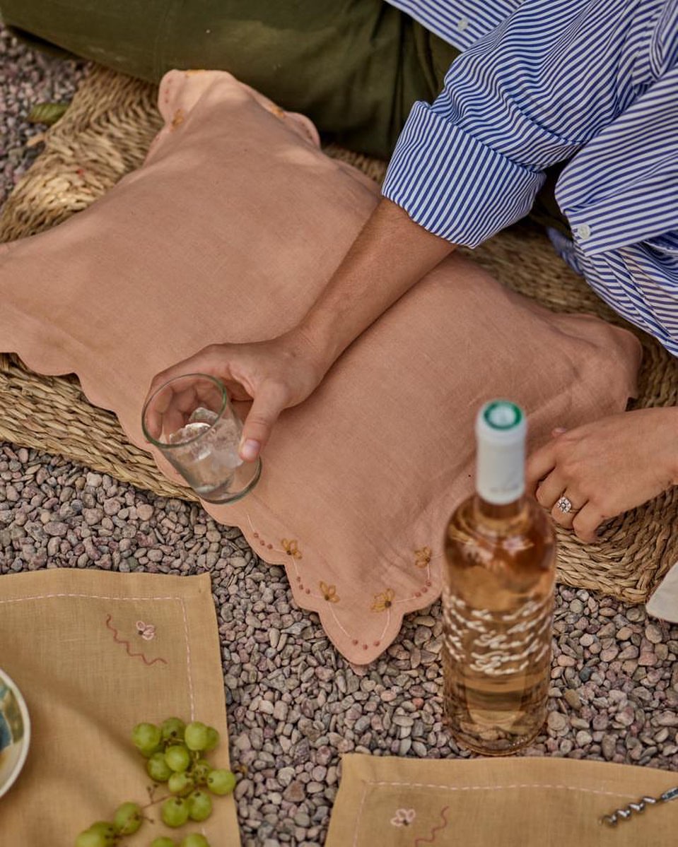 Outdoor picnic scene featuring a pillow, wine bottle, glass, grapes, and two people enjoying a casual meal on a gravel surface.