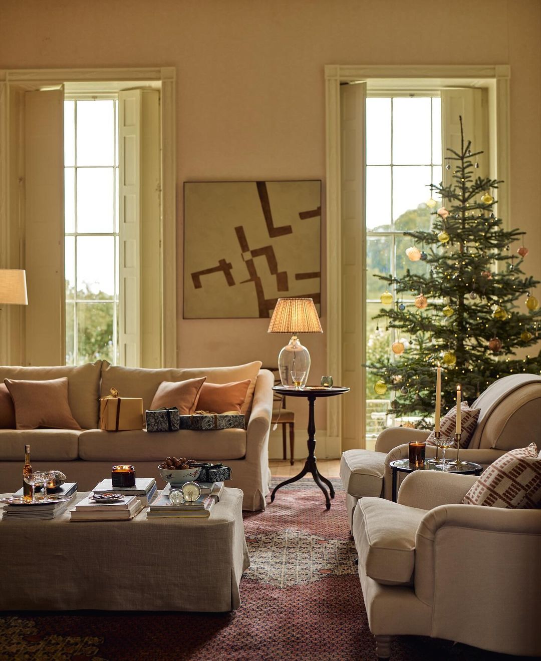 Living room interior with a Christmas tree near a window, sofa, armchair, ottoman, table, and indoor decorations in a warm, inviting setting.