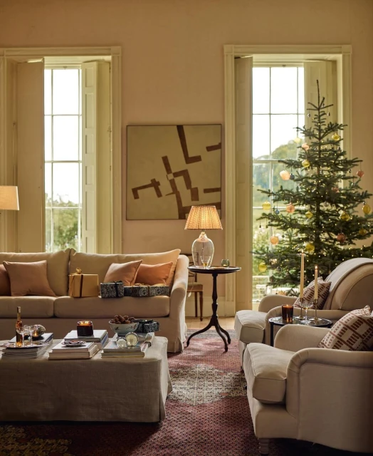 Living room interior with a Christmas tree near a window, sofa, armchair, ottoman, table, and indoor decorations in a warm, inviting setting.