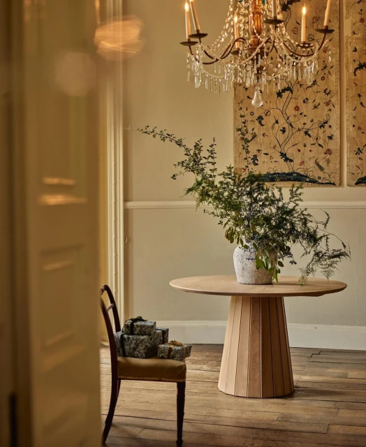 Interior view of a round wooden table topped with a vase of greenery, a crystal chandelier hangs above, and gifts sit on a chair.