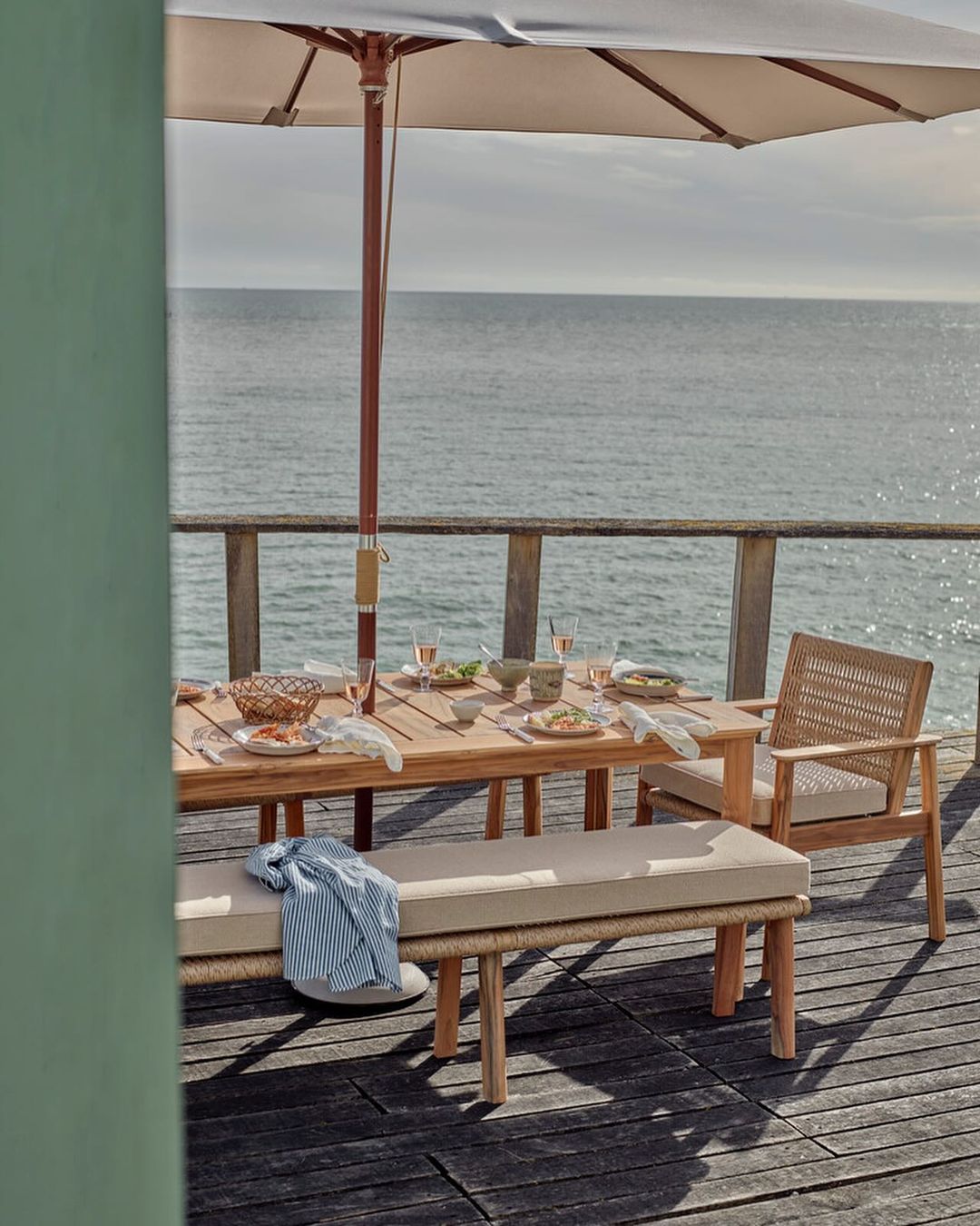 Outdoor dining table setup with light wood furniture and tableware under a parasol with ocean view on a sunny day.