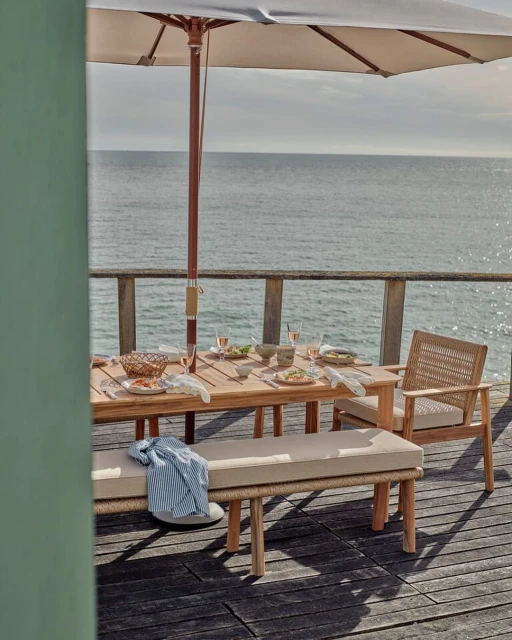 Outdoor dining table setup with light wood furniture and tableware under a parasol with ocean view on a sunny day.