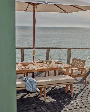 Outdoor dining table setup with light wood furniture and tableware under a parasol with ocean view on a sunny day.