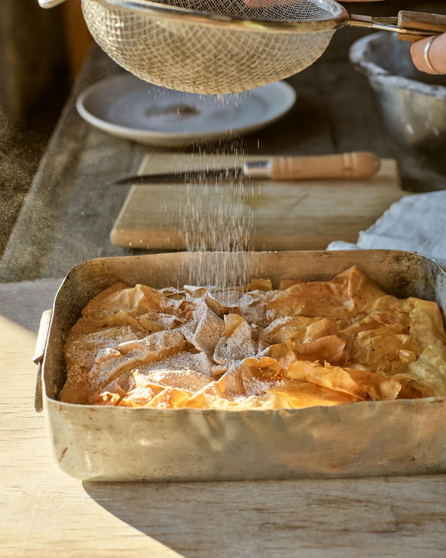 Sprinkling powdered sugar on baked good in metal pan with sifter, wooden table, and hands in soft light.