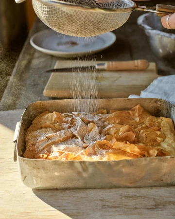 Sprinkling powdered sugar on baked good in metal pan with sifter, wooden table, and hands in soft light.