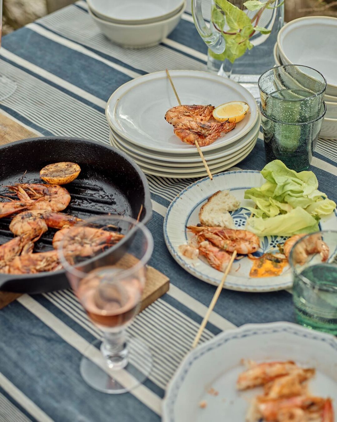 Grilled shrimp skewers with lemon wedges and a glass of rosé wine on a blue and white striped tablecloth, outdoors, for food photography.