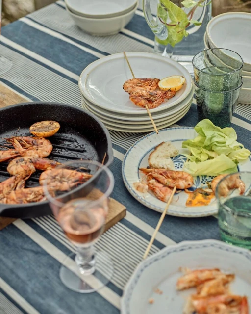Grilled shrimp skewers with lemon wedges and a glass of rosé wine on a blue and white striped tablecloth, outdoors, for food photography.