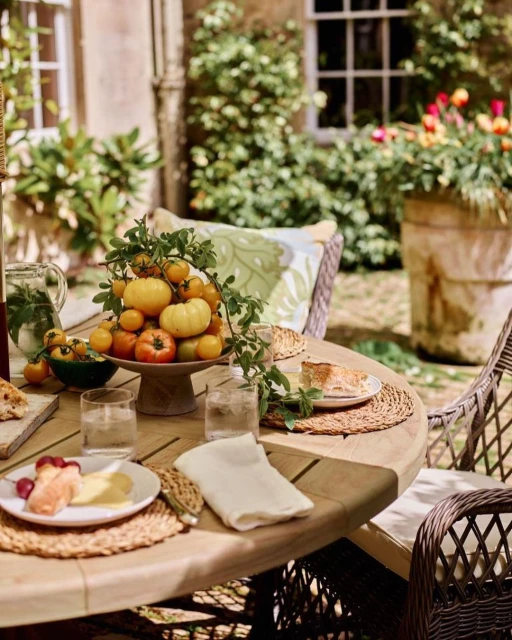 An outdoor dining setting featuring a wooden table with place settings, fruit, and cheese, surrounded by lush greenery and stonework.