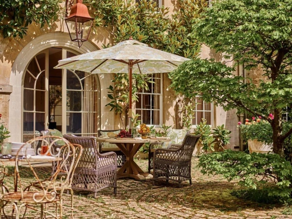 Outdoor dining area with a round wooden table, woven chairs, and a floral umbrella in a courtyard setting with lush greenery.
