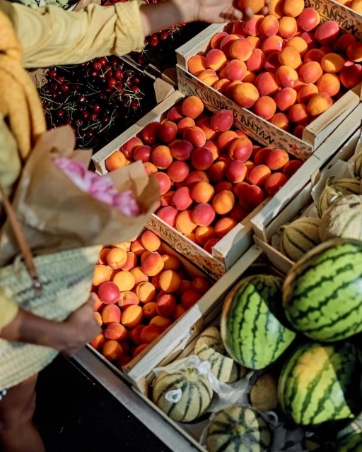 A high-angle view displays several wooden crates filled with apricots, cherries, and watermelons at a fresh food market.
