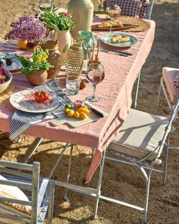 Outdoor dining table set with a pink tablecloth, various dishes, flowers, drinks, and chairs on a gravel surface in daylight.