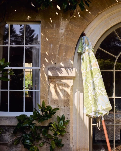 Exterior shot featuring an umbrella with a green and white floral pattern, positioned near arched windows and a stone wall with greenery.