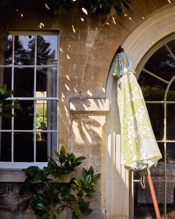 Exterior shot featuring an umbrella with a green and white floral pattern, positioned near arched windows and a stone wall with greenery.