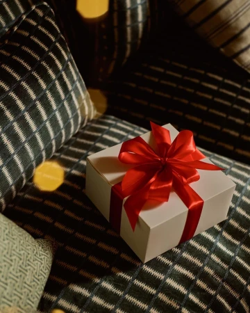 Festive white gift box tied with a bright red satin ribbon, resting on a patterned couch cushion under soft, warm lighting.