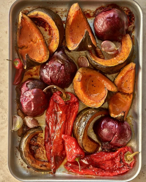 Overhead shot of roasted vegetables, including butternut squash, red onion, red bell pepper, and garlic, arranged on a metal baking sheet.