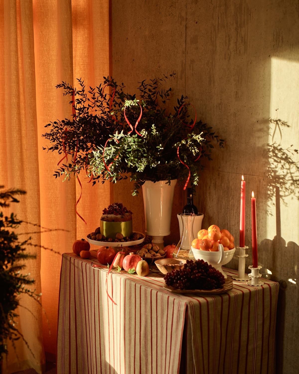 Festive table setting with cake, fruit, and candles, illuminated by warm light against an orange curtain and textured wall backdrop.