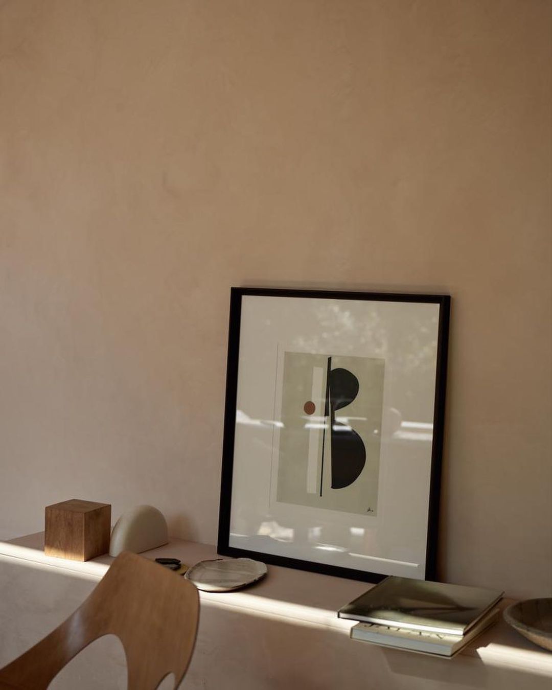Artwork featuring geometric shapes in a minimalist composition with a black frame, resting on a table near decorative objects and a chair.