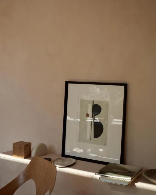 Artwork featuring geometric shapes in a minimalist composition with a black frame, resting on a table near decorative objects and a chair.