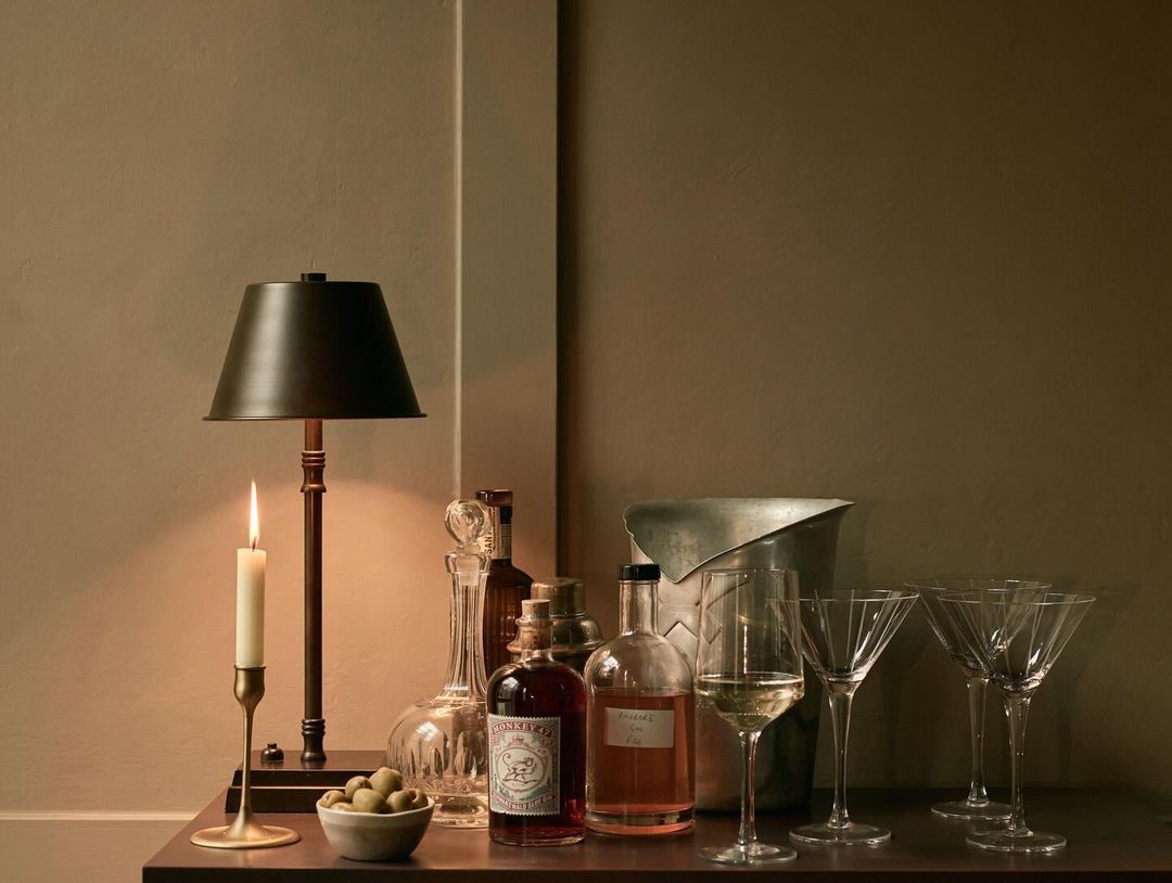 Still life shot of a bar area featuring drinks, glasses, a table lamp, and a lit candle, set against a neutral color wall.